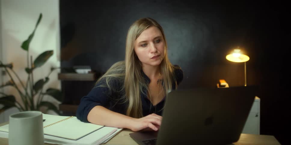 Blonde woman with black shirt sitting at desk uses laptop and gets surprised in front of black wall