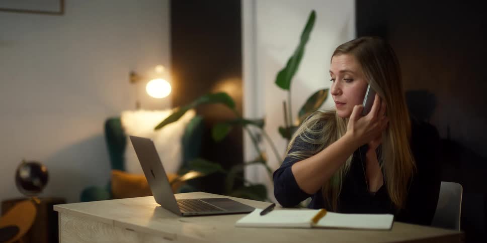 Blonde woman with black shirt sitting at desk talks on smartphone while using laptop