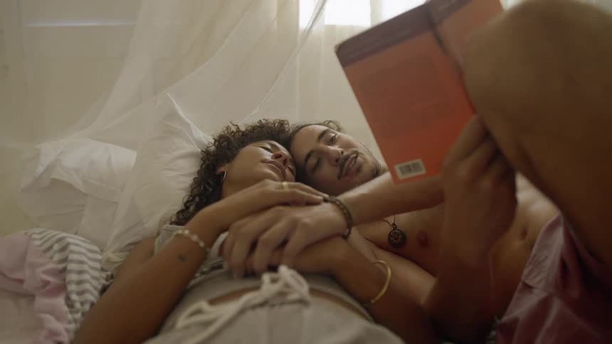 A man reads a book aloud while lying in bed with his resting wife