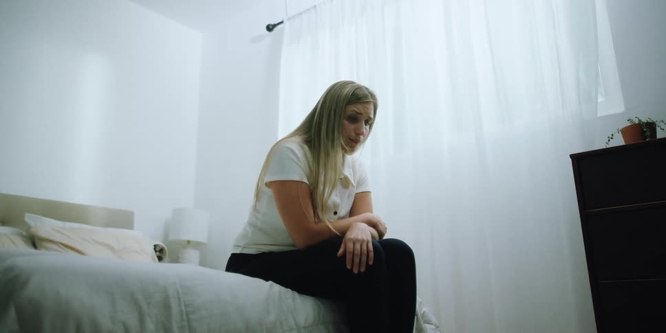 Young sad crying depressed woman sitting on bed inside a white bedroom