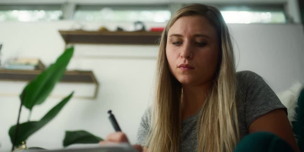 A woman with long blonde hair writing in a notebook while sitting in a chair