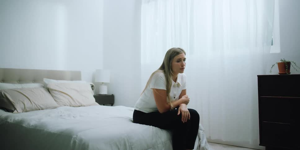 A woman sits at the end of her bed crying and looking distraught