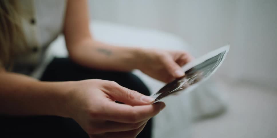 A woman in a room sitting on a bed looking at a picture of an ultrasound with a wall in the background