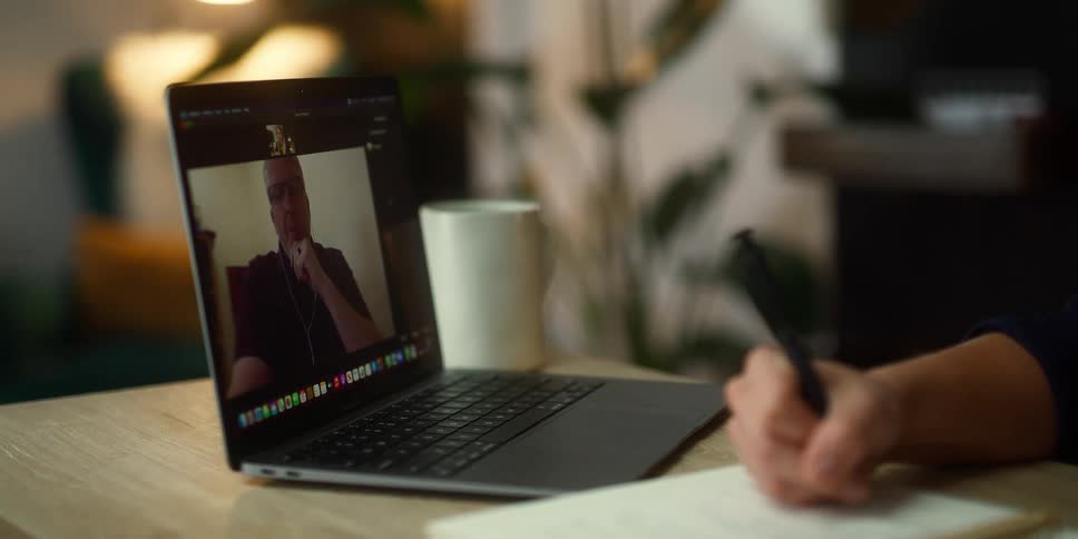 A person sits at a table writing in a notebook with a laptop open for a video call
