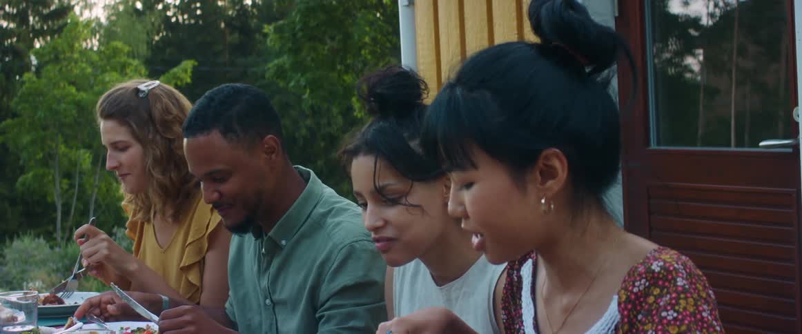 A group of friends are having a meal outdoors at a picnic table in a backyard