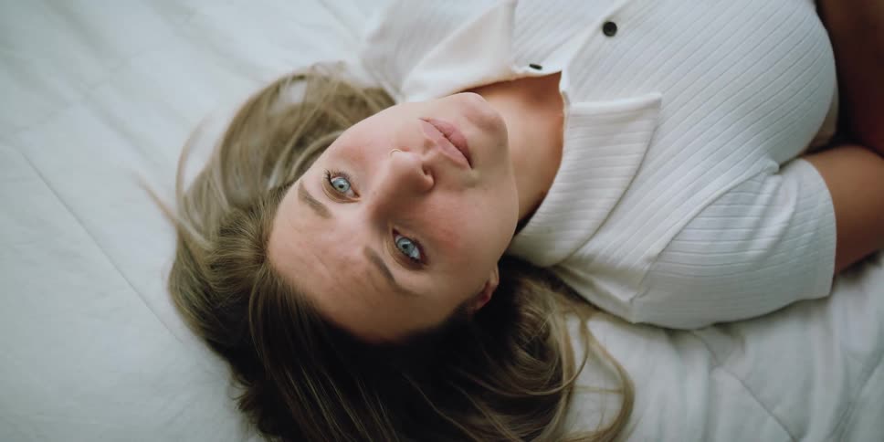 A young woman flops back on a bed with a worried look on her face