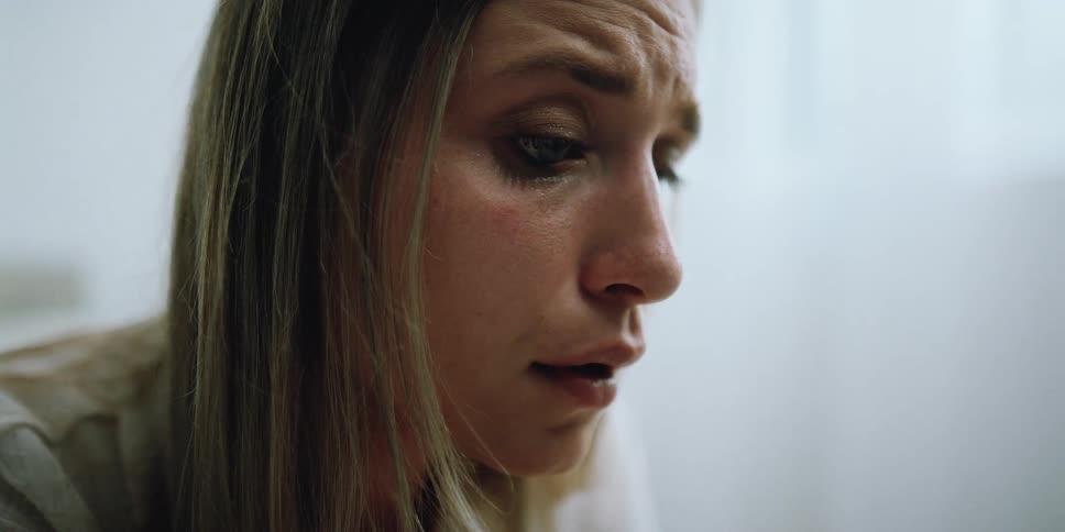 A woman sitting on a bed in a room with tears in her eyes with a wall and window in the background
