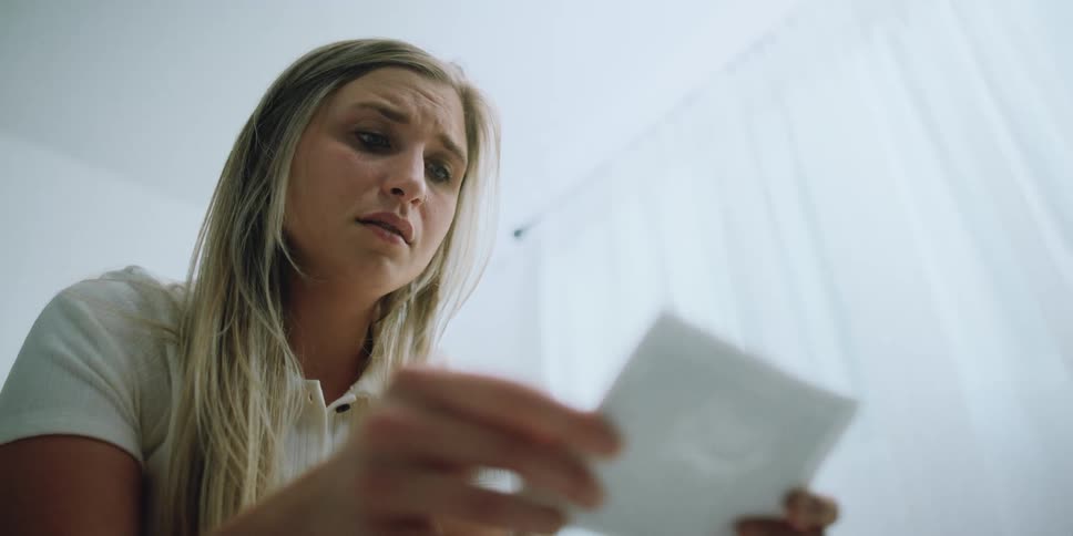 Pregnant blonde woman with white shirt looks down at x ray pregnancy photo getting emotional