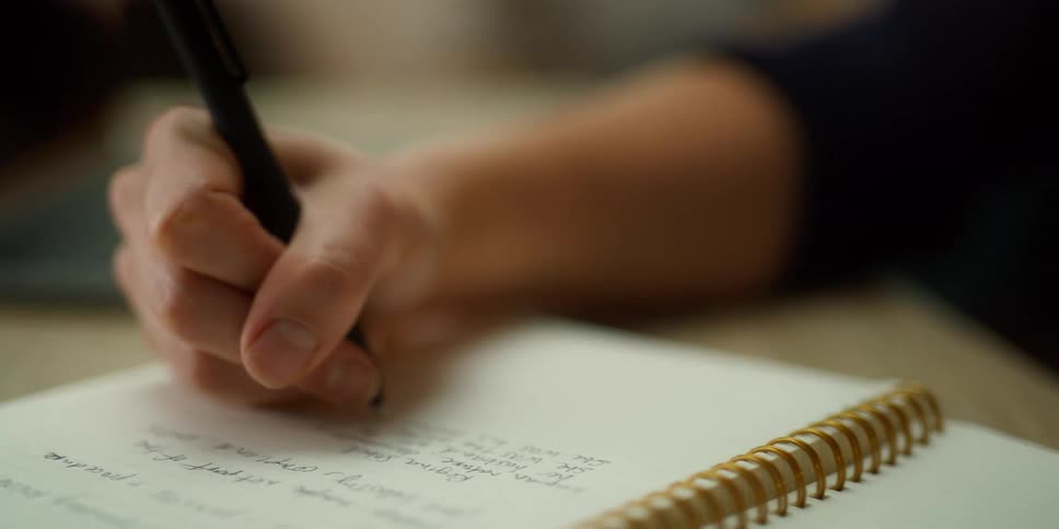 A young person is holding a pen and writing in a spiral notebook