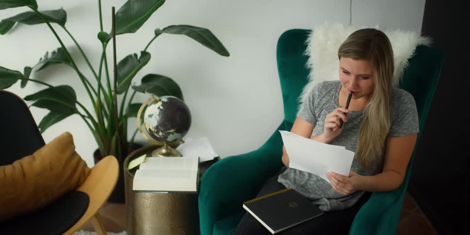 A woman in a room sitting on a couch holding a pen with a wall in the background