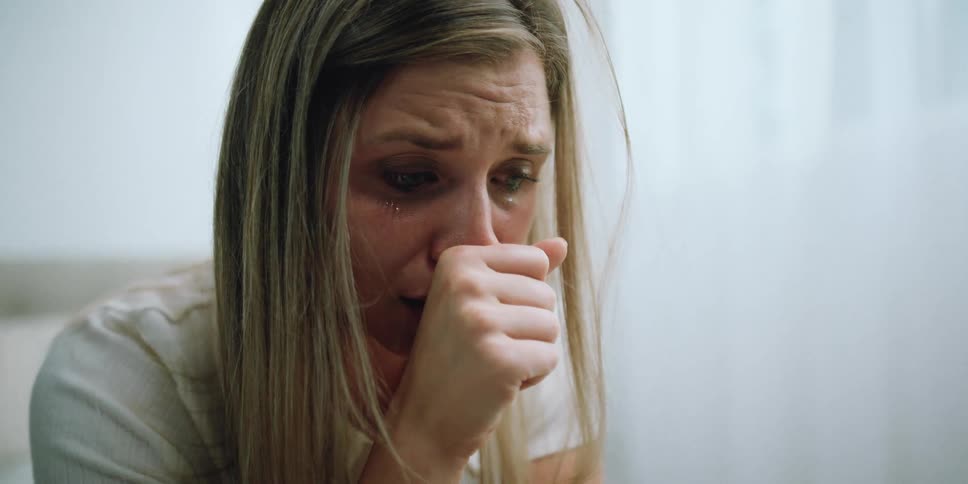 A woman in a room sitting on a bed with tears in her eyes with a wall and window in the background