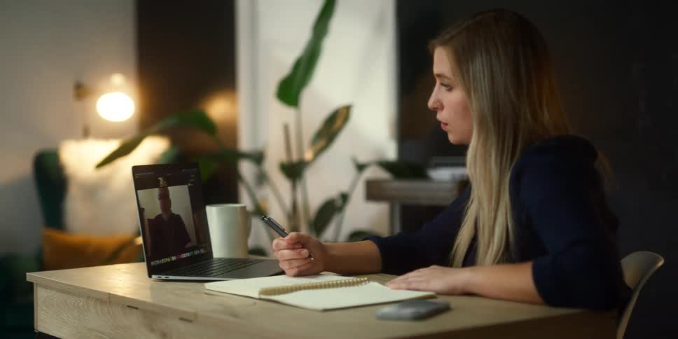 Blonde woman with black sweater sits at desk makinga video call and taking notes