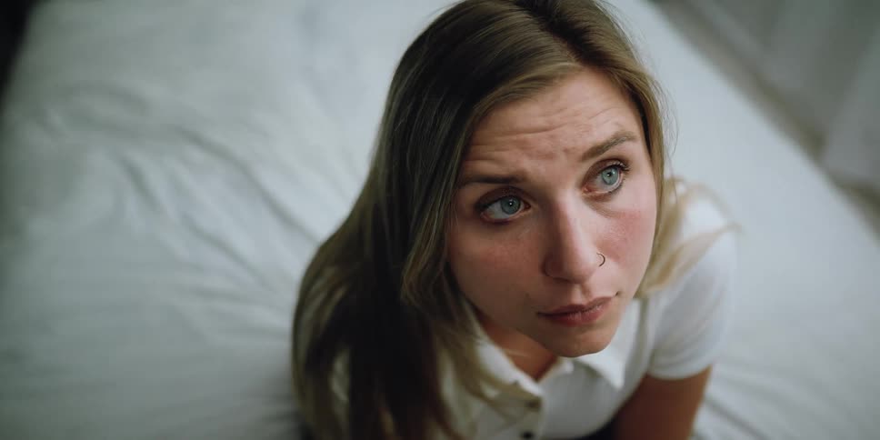 Blonde woman with blue eyes and nose ring sits on bed and looking up at ceiling pondering
