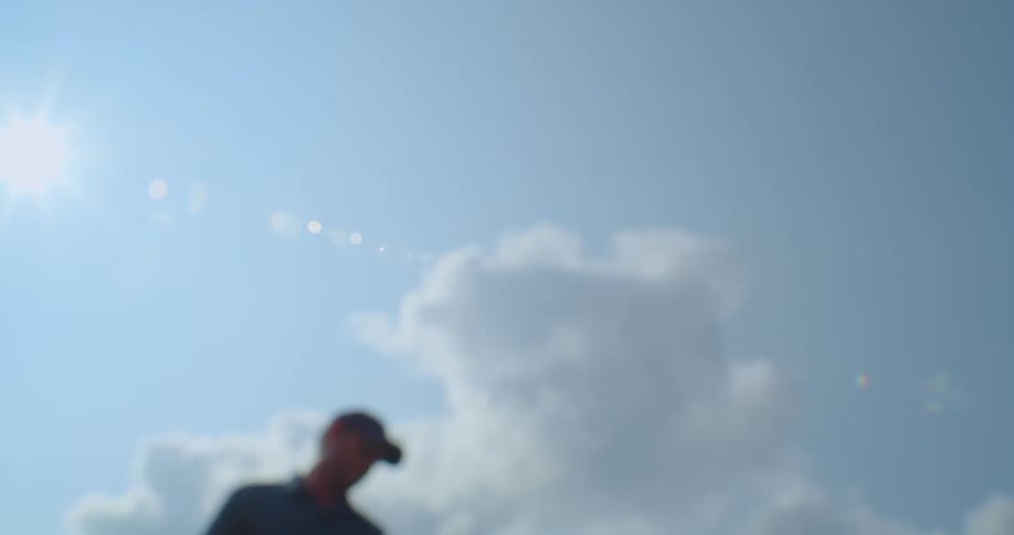 A man with a tennis racket picking up a tennis ball and hitting it with the racket with the sky and clouds in the background