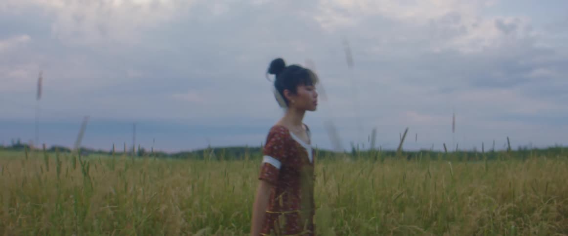 A young woman runs her hand along stalks of grass as she walks through a field of tall grass on a cloudy day