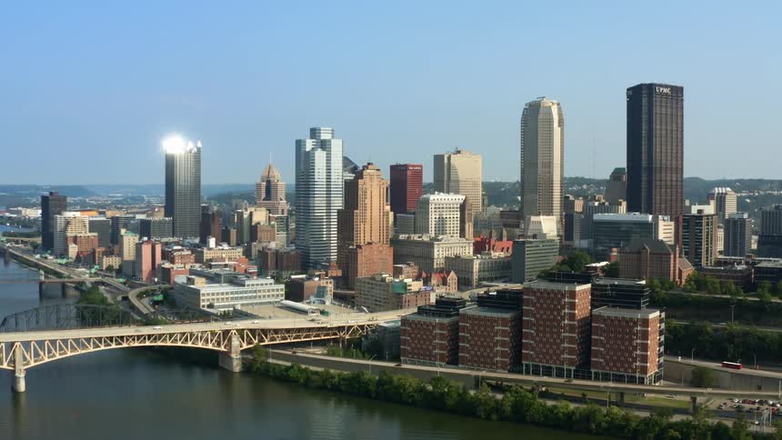 A city skyline with buildings and skyscrapers with a bridge next to a river with the sky and clouds in the background