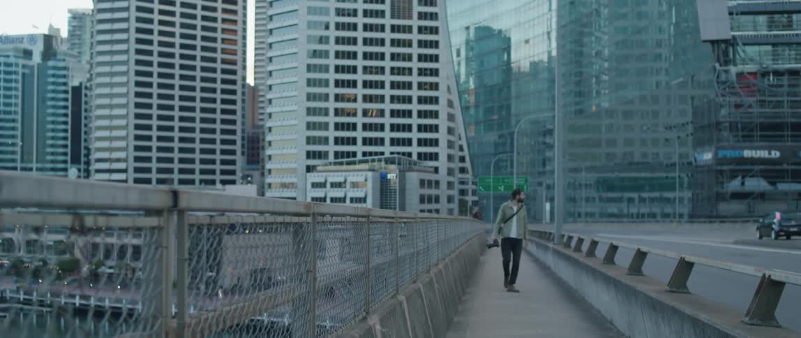 Photographer with beard crosses bridge with tall buildings in the background