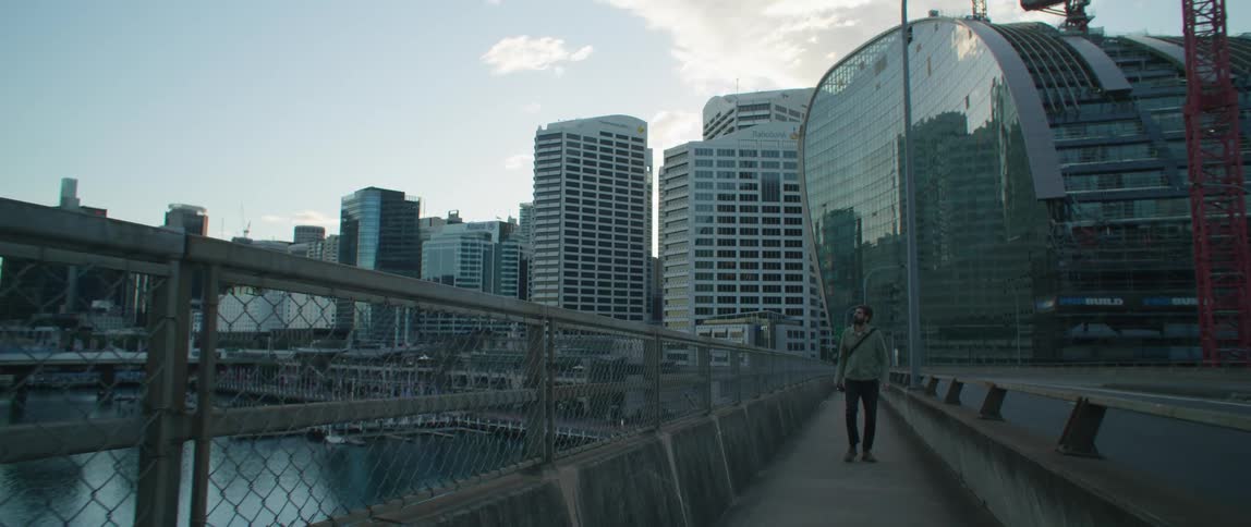A man walking on a walkway with a fence next to a marina with buildings the sky and clouds in the background
