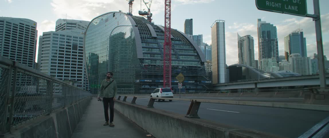 A man walking on a sidewalk in front of large buildings in a city looks around as cars drive past him