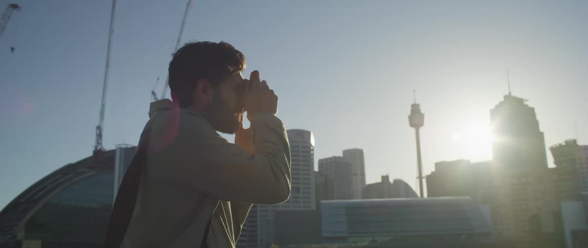A man stands in front of office buildings and construction cranes in a city and takes a photo