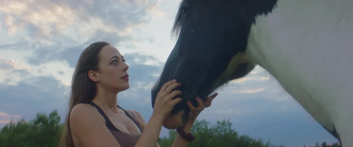 Woman with brown hair and sleeveless shirt caresses horses head at dusk