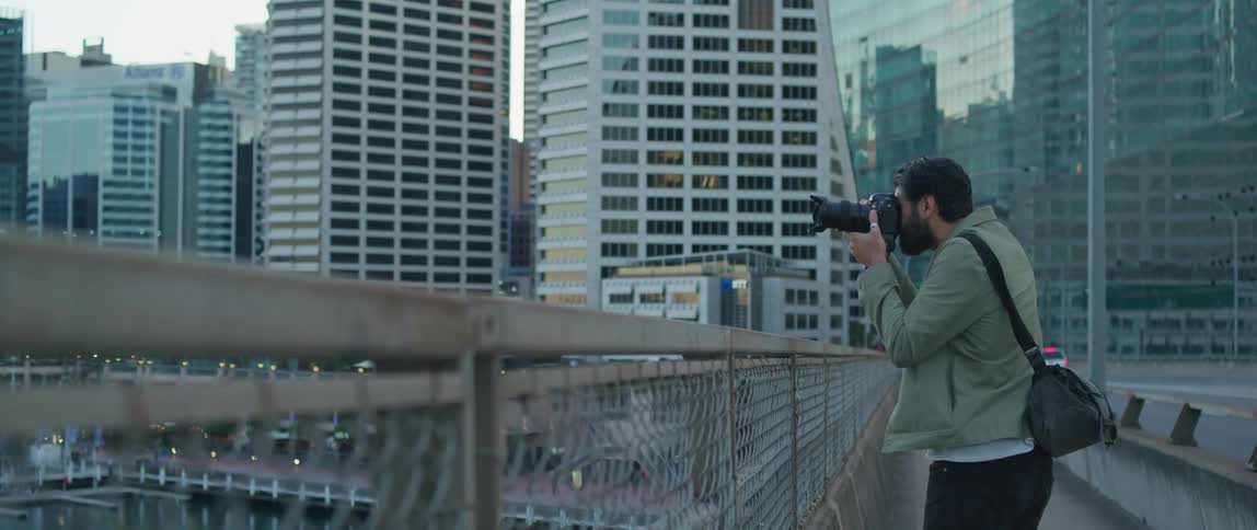A man with a camera standing on a bridge and taking a picture with buildings the sky and clouds in the background