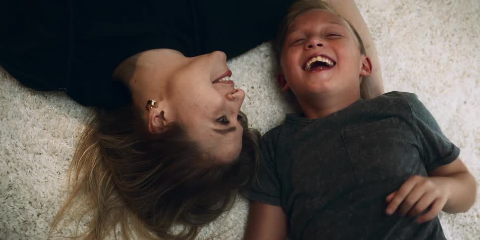 A woman and a boy laugh as they lie next to one another on a carpet indoors