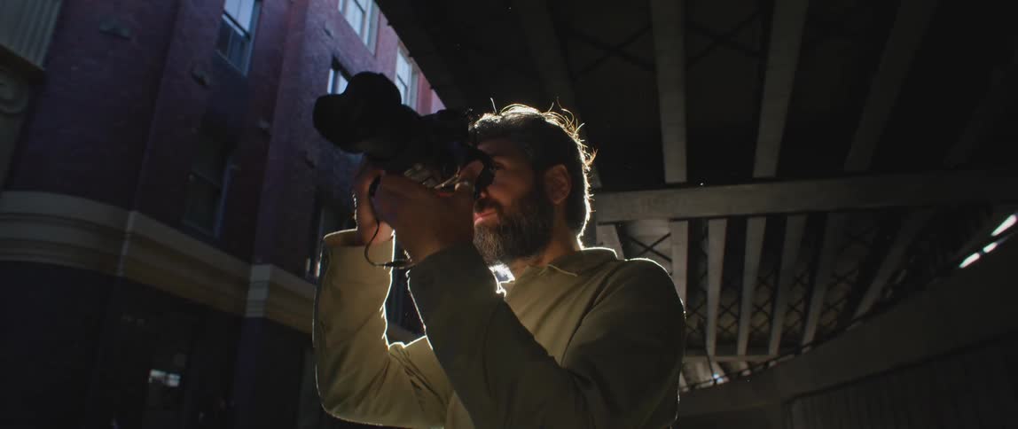 A man with a camera underneath an overpass with a building and the sun in the background