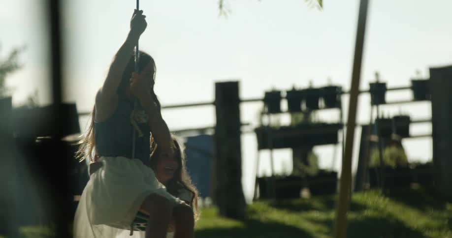 One girl on a swing while another girl pushes her with potted plants the sky and clouds in the background