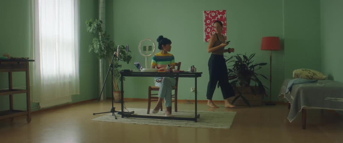 A woman putting on makeup while another woman picks up things behind her