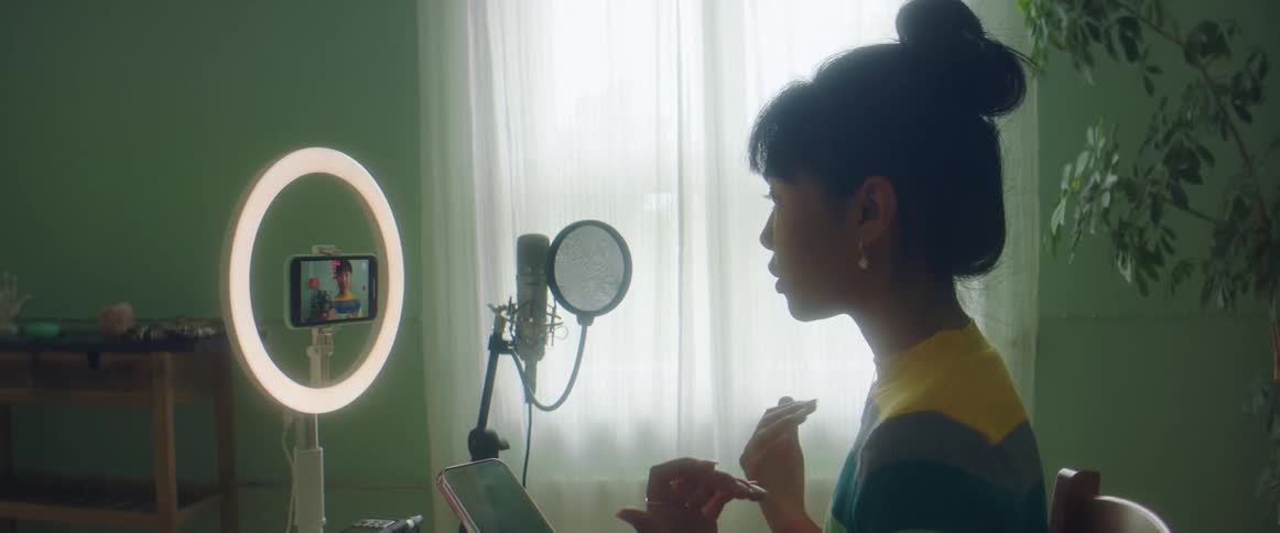 A woman in a room videocasting with a smart phone with a window and a wall in the background