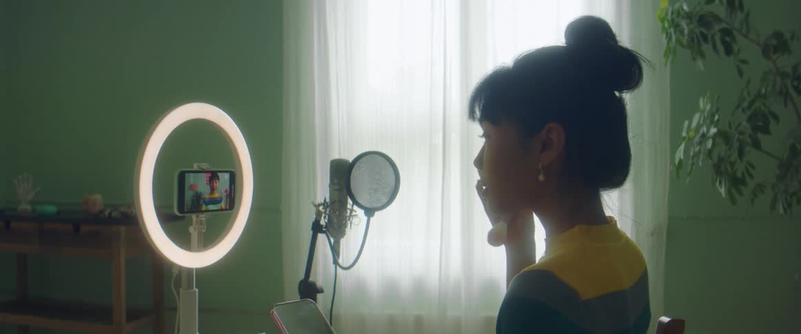 A young woman applies makeup to her face while recording herself with a smartphone mounted on a stand