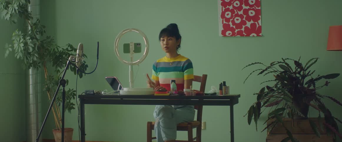 A woman sits at a table applying makeup and recording a product demonstration video on her smartphone