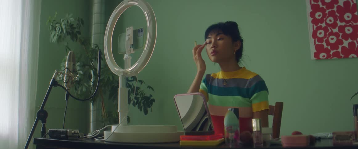 A woman in a room sitting on a chair applying makeup to her face with a brush with a wall and window in the background