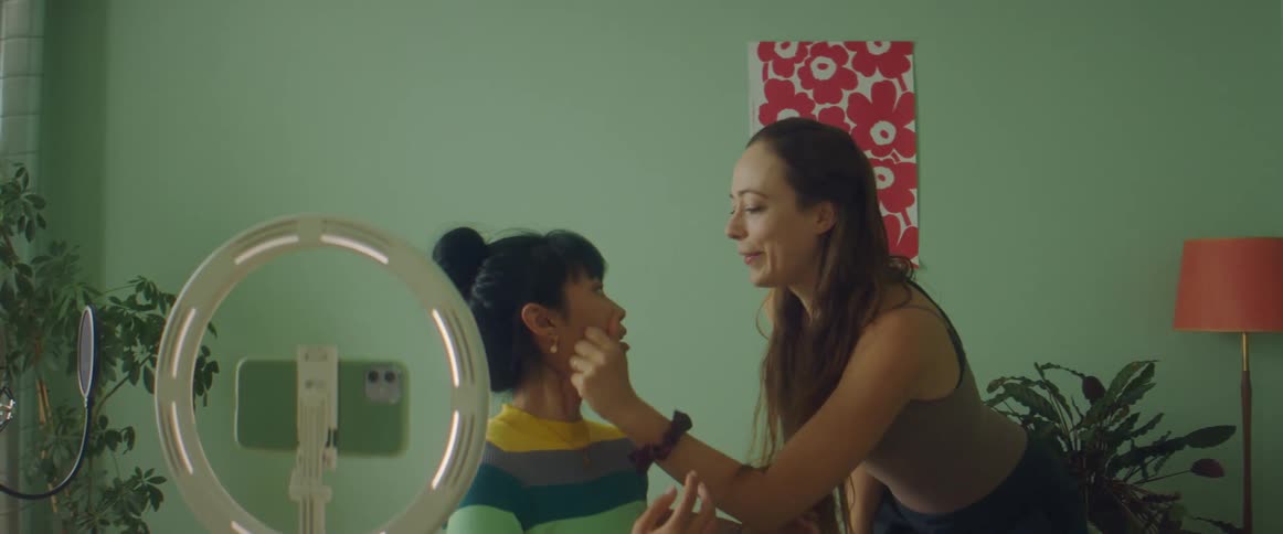 A woman puts on makeup in front of a ring light and smart phone and another woman comes and squeezes her face playfully