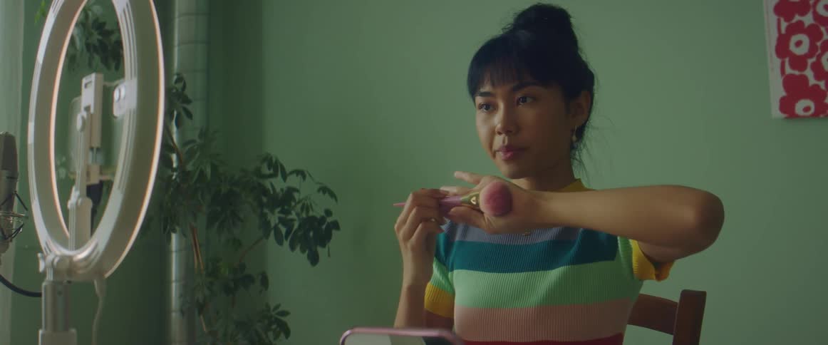 Woman with colorful shirt sits at table and puts on makeup while filming herself on smartphone