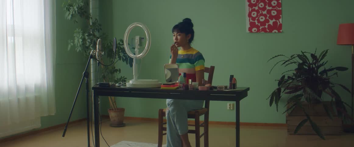 Woman sits at table putting on makeup and smiling into camera near microphone stand