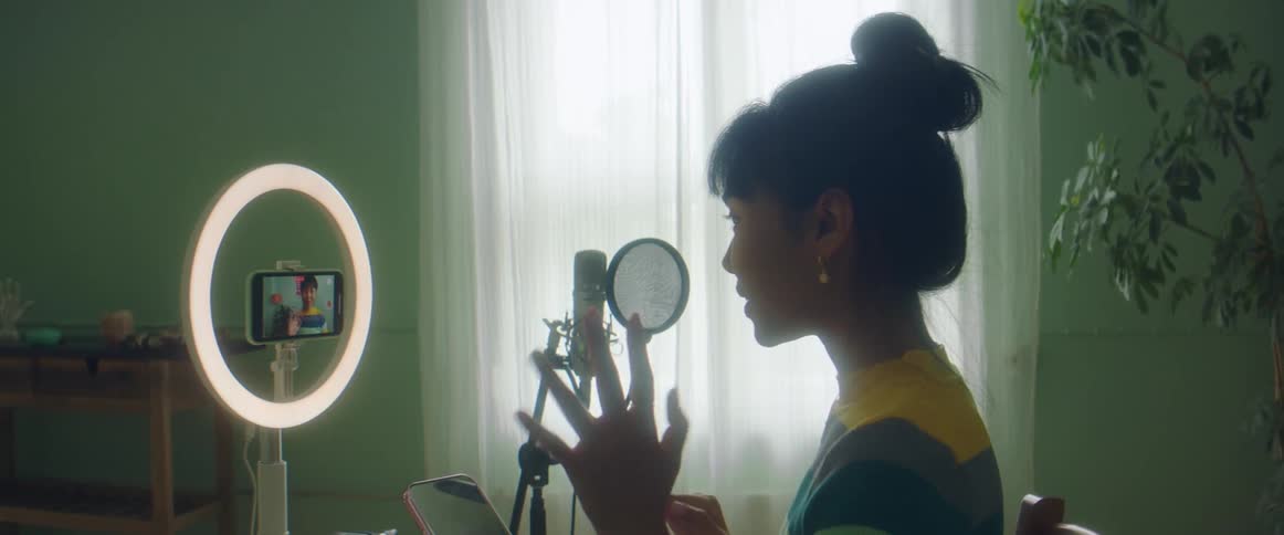 A young woman records herself with a smartphone mounted in an illuminated stand as she speaks