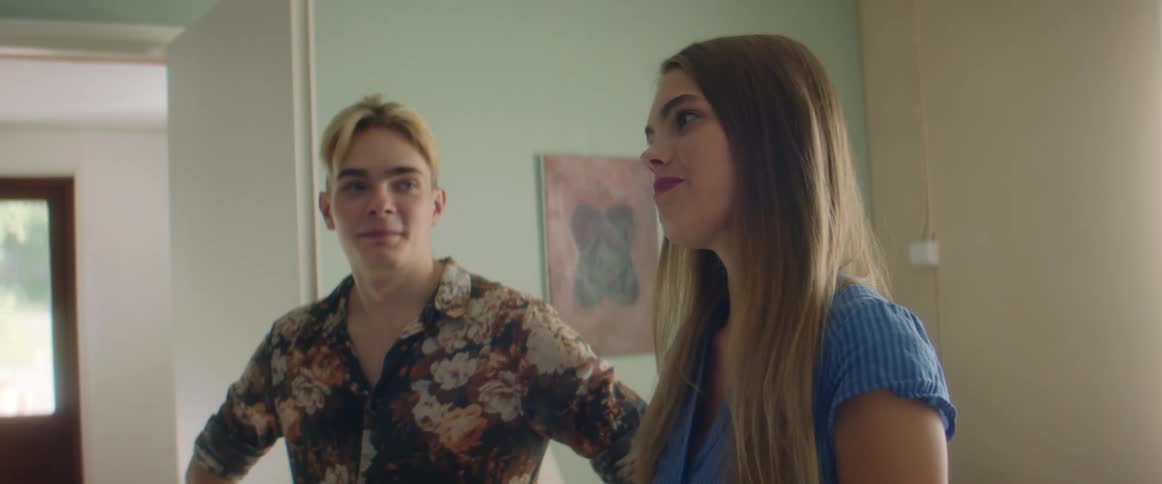 A teenage boy and girl stand in a house and talk with others