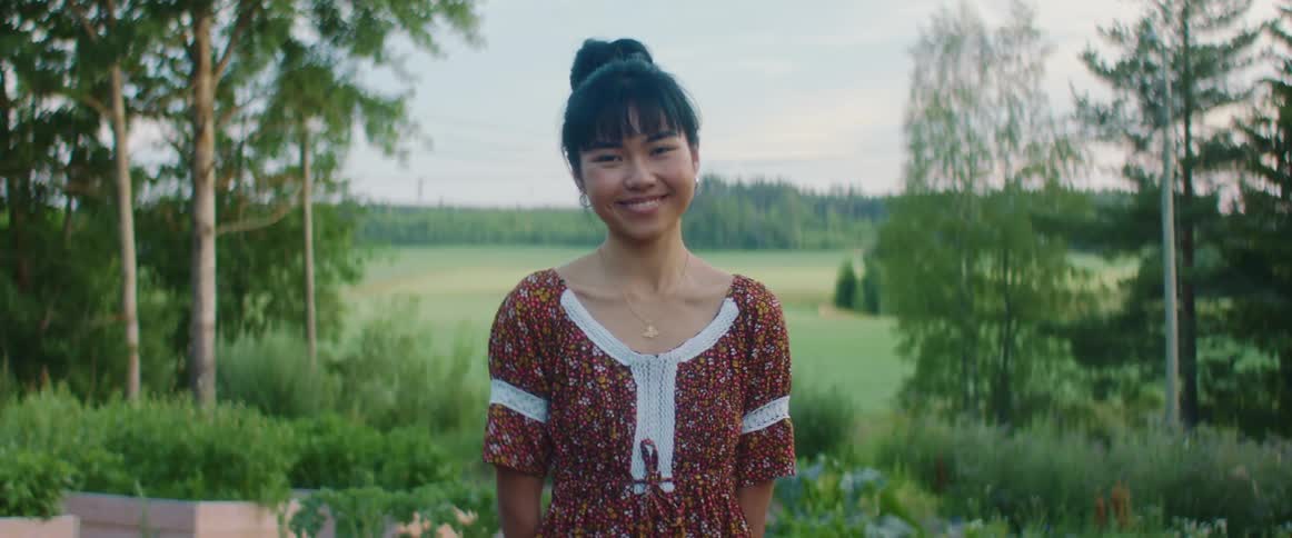 A young woman wearing a dress is smiling and standing outdoors near a garden with a large field and trees in the background