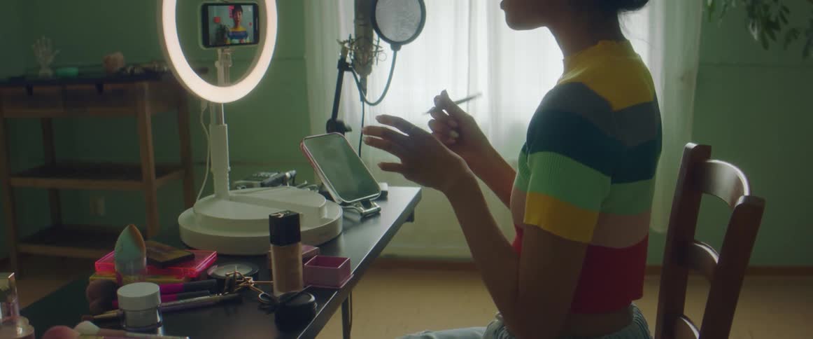 A woman in a room sitting on a chair applying makeup with a brush with a window and a wall in the background