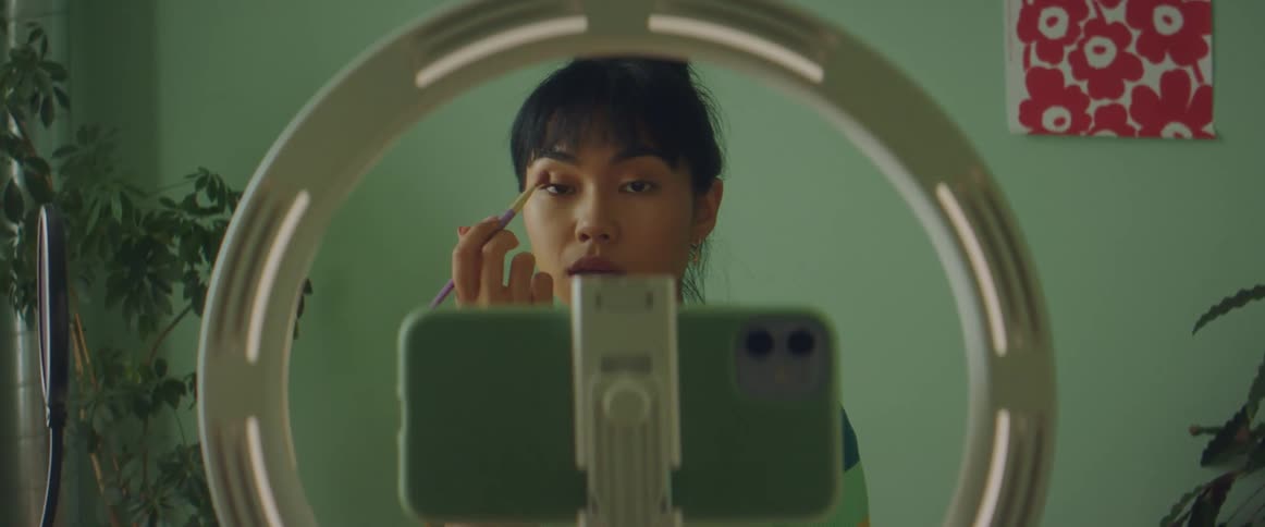A teenage girl sits in front of her cell phone and records herself as she puts on make up