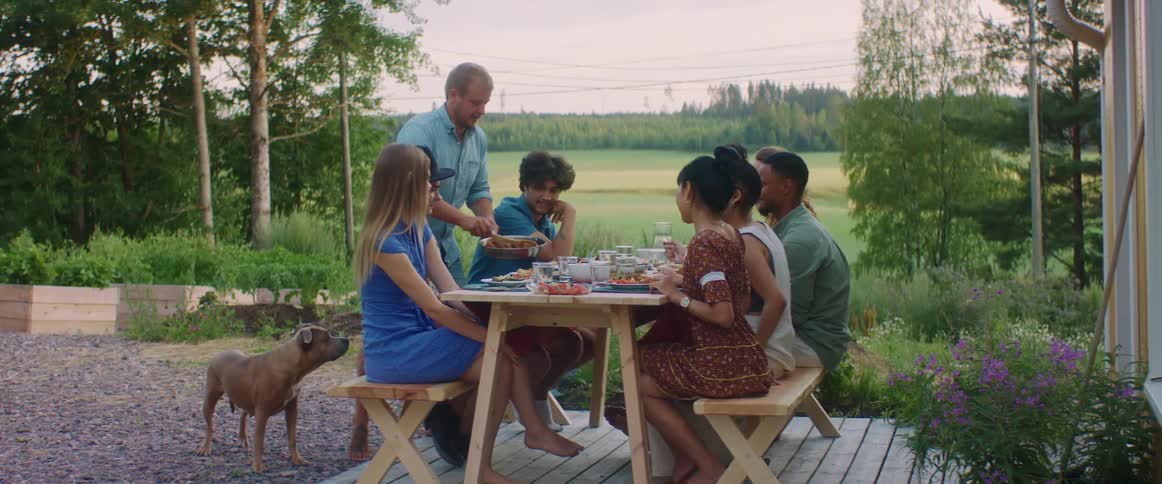 A man serves food to a group of friends sitting at a picnic table in the yard