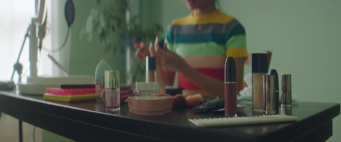 Woman with colorful shirt sits at desk with cosmetics and films herself putting on makeup