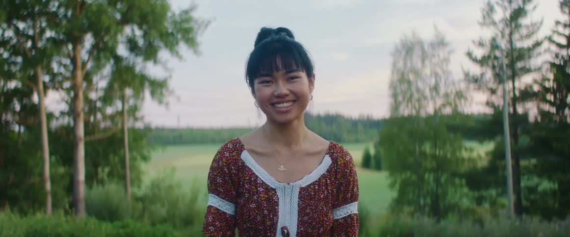 A woman standing staring and smiling with trees a field the sky and clouds in the background