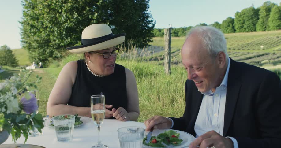 A man and woman sit at a table on a vineyard and talk as they enjoy a meal with wine