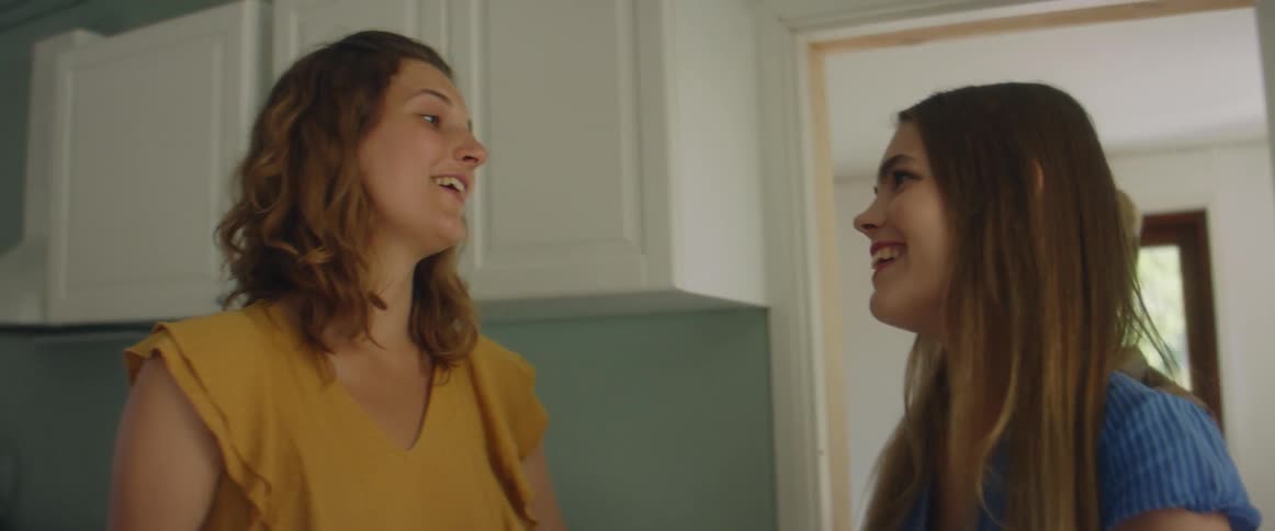 A woman opens her house door to welcome two of her friends and she hugs them as they enter