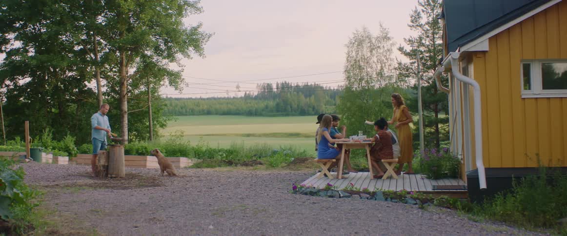 A man is cooking on a grill while a woman sets dishes down on a table with guests