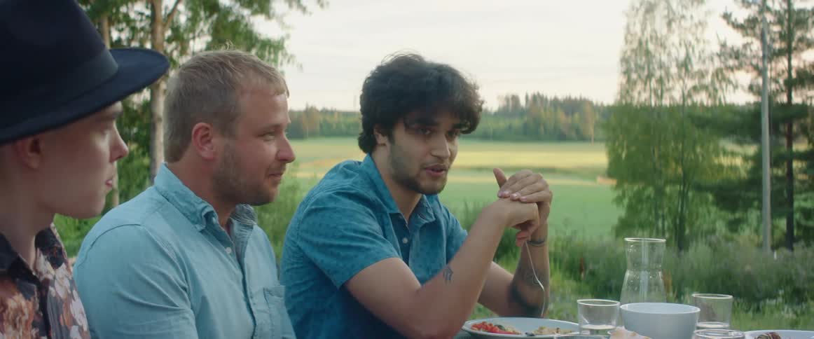 Friends sit around outside at picnic table talking and eating with field and forest in background