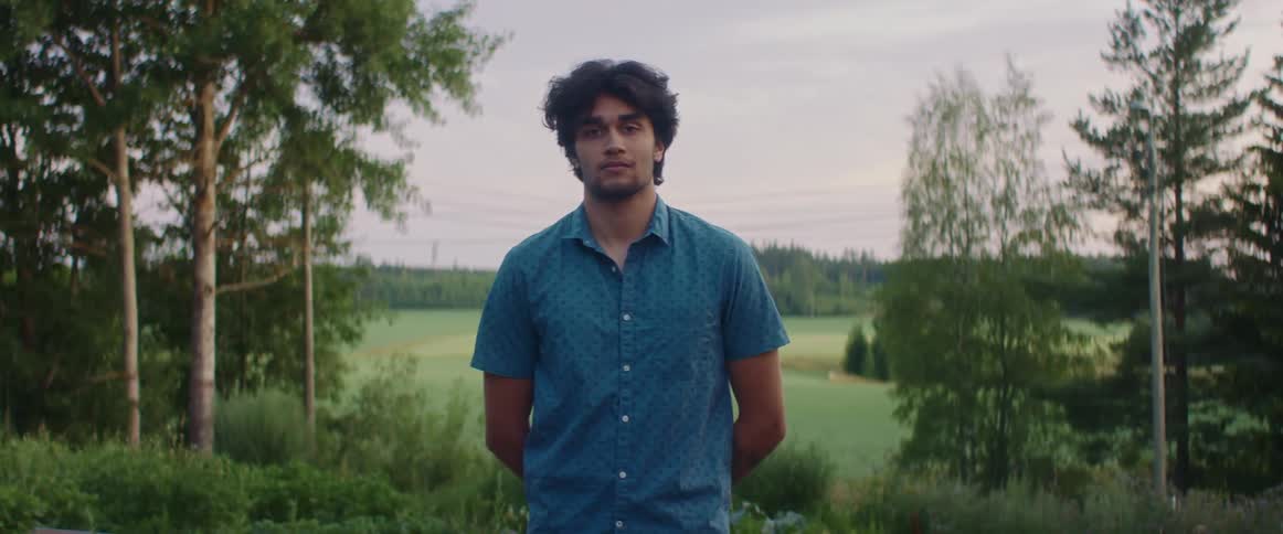 A young man stares into the camera while standing in the countryside outdoors in the evening
